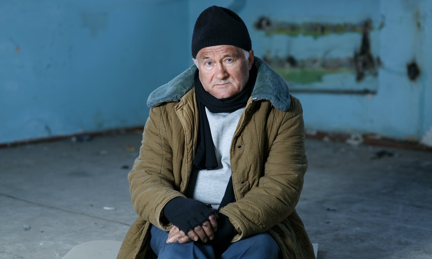 An older man sits bundled up in a dilapidated space, looking cold and in need of assistance.