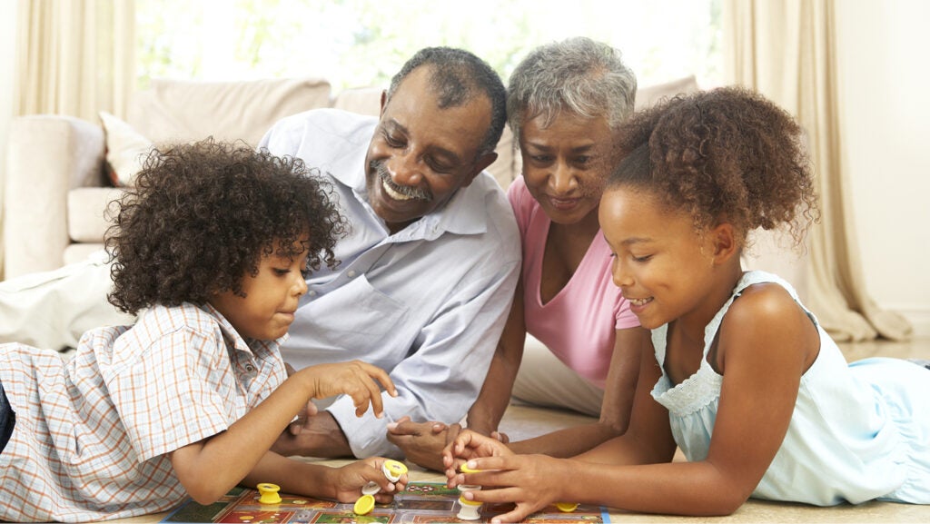 Grandparents and two grandchildren, a boy and a girl, lie on the living room floor, playing a board game. They're all smiling and happy.