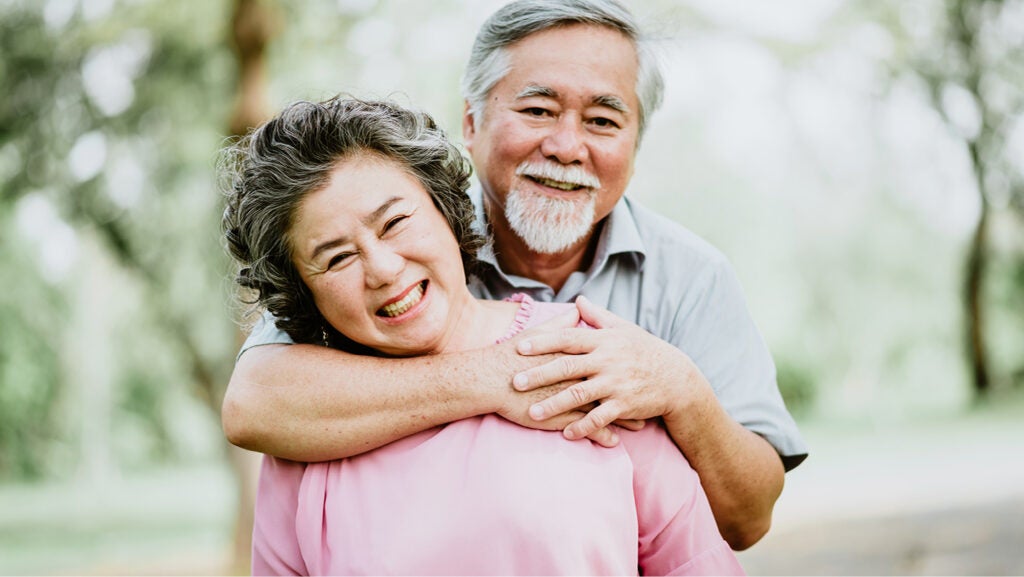 A smiling older couple stand together. He has a goatee and his arms around the woman, who wears a pink top.
