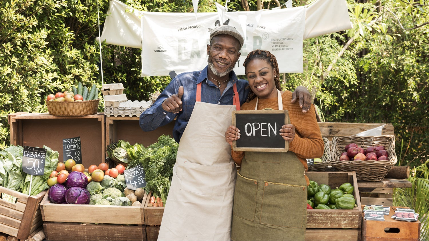 A happy couple in aprons stands in front of a produce stand. He has his arm over her shoulder and is giving a thumbs up while she holds a small chalkboard reading "Open."