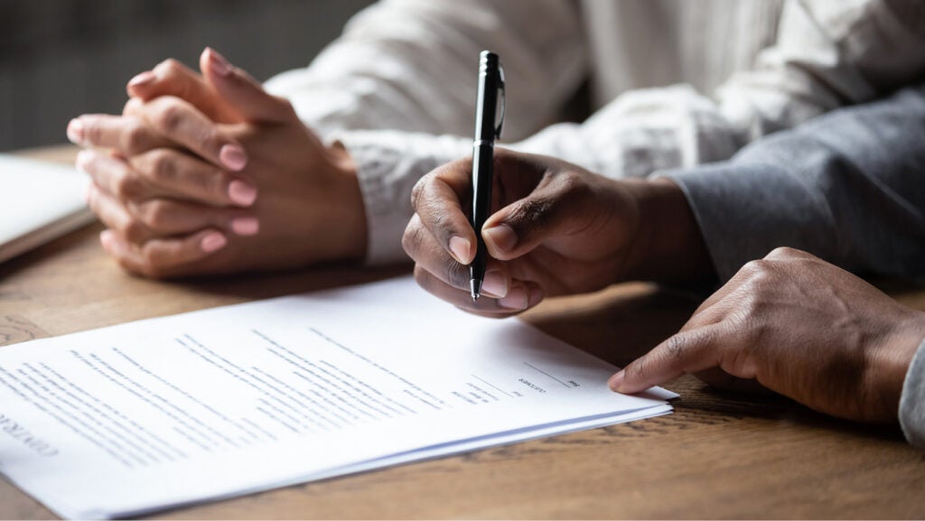A closeup of the hands of two people. One holds a pen and prepares to sign paperwork. The other has her hands folded on the table.