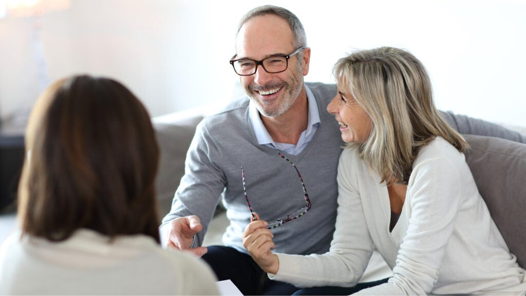 A smiling older couple sit on a couch. They are wearing v neck sweaters and she holds her glasses in her right hand. They're speaking with an investment advisor.