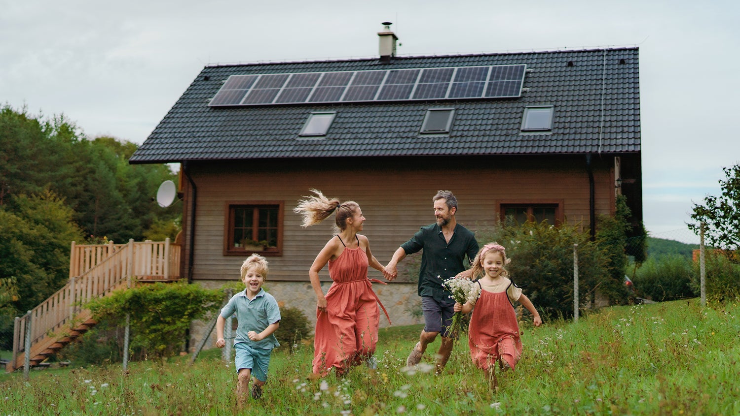 A family of four run in a green field in front of a wooden house with solar panels on the roof. The parents hold hands and the boy and girl run ahead of them, the girl with flowers in her hand.