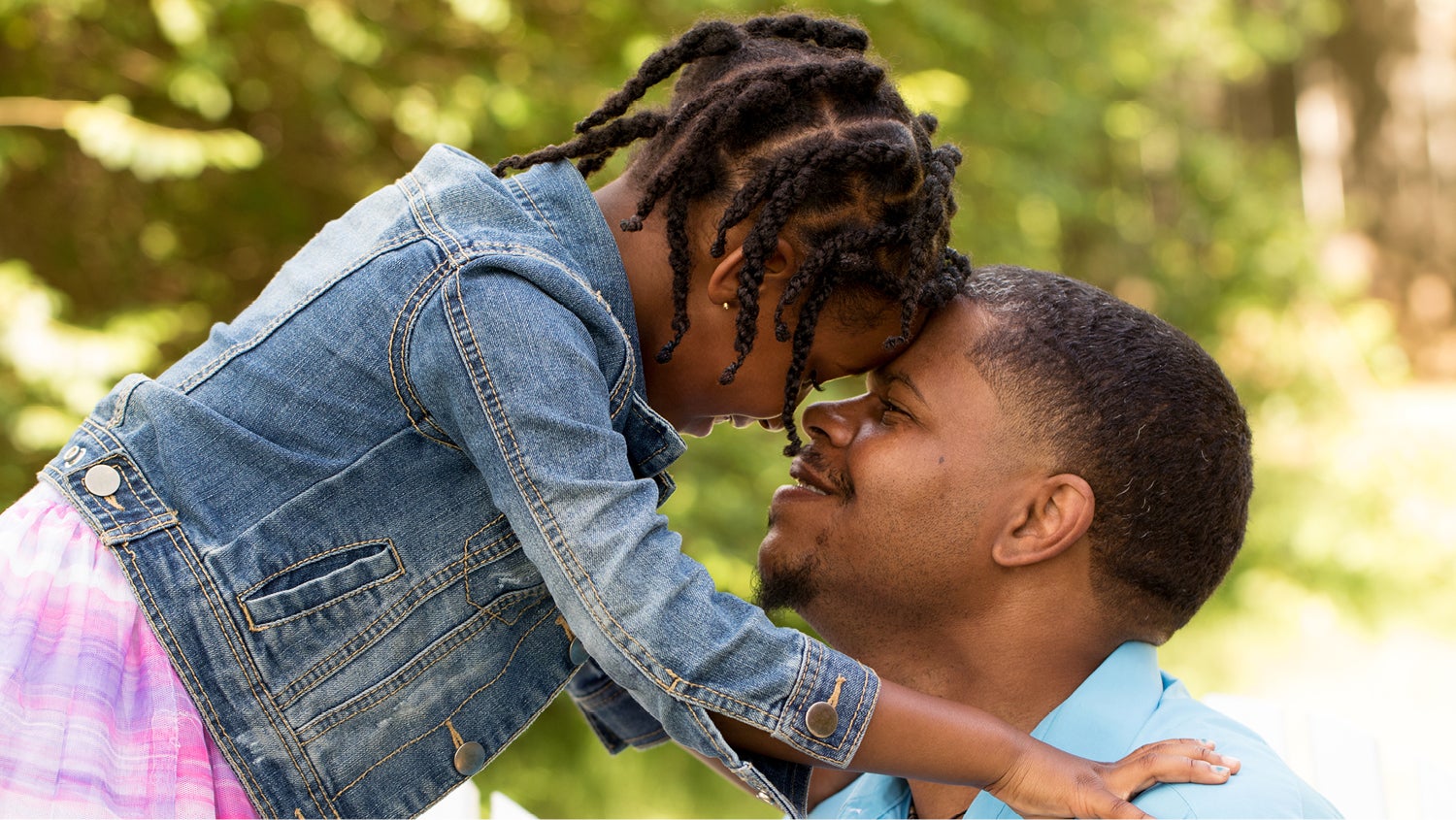 A little girl with braids in her hair wearing a jean jacket puts her forehead against her father's forehead. They are smiling at each other.