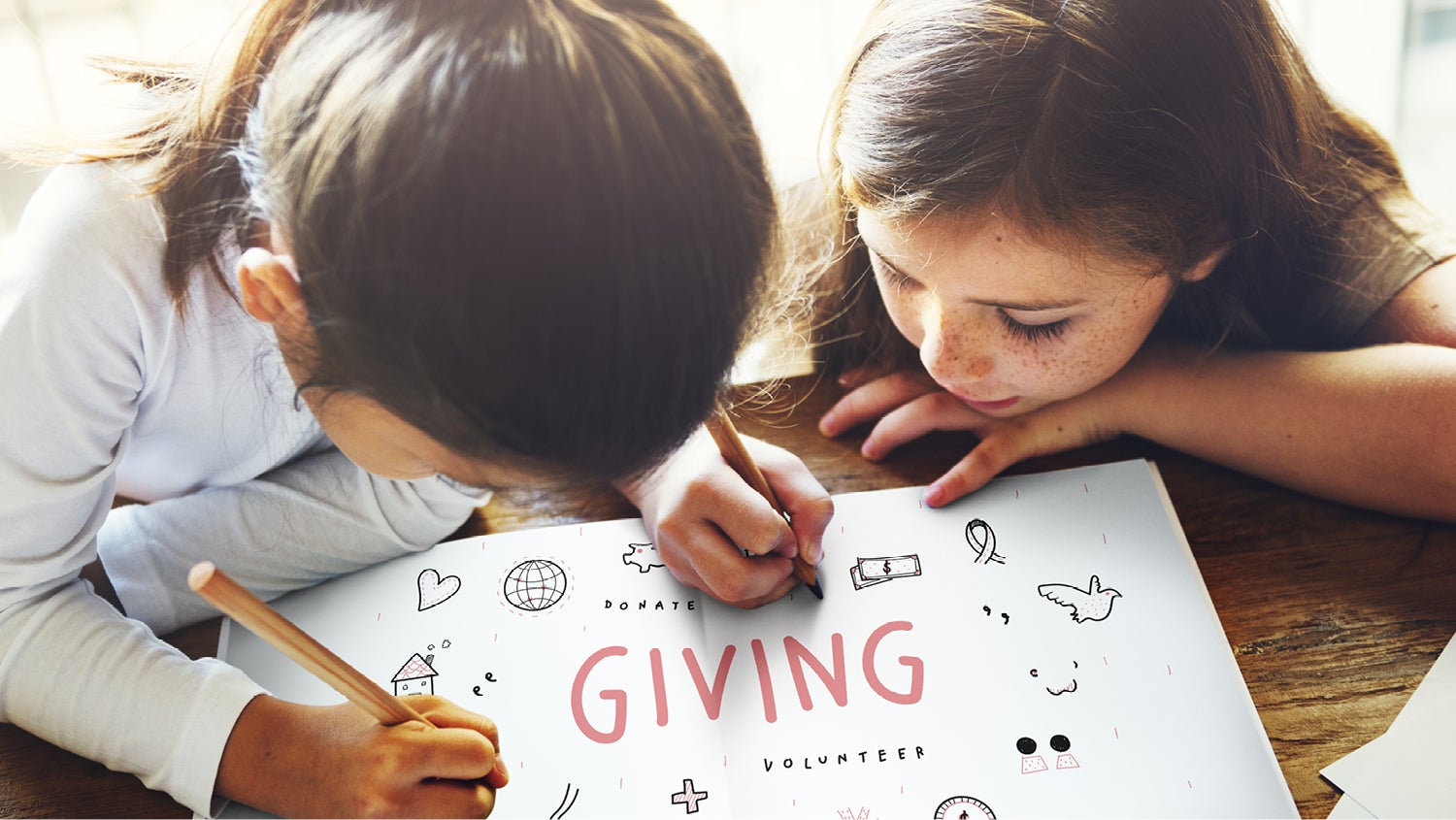 Two little girls lie on their stomachs, drawing on a sheet of white paper that says "Giving" and "volunteer" on it.